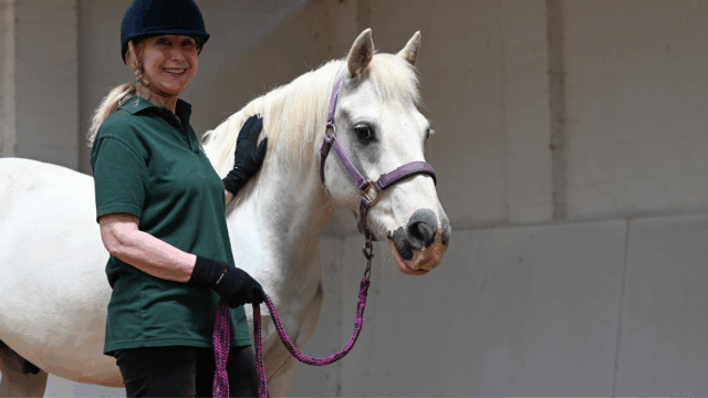 Image of an adult woman wearing a black riding helmet and a green polo shirt, standing next to a grey pony, named Toptails. Candy a volunteer is smiling and has her gloved hand resting on the pony's neck. The pony has a light-colored mane and is wearing a purple halter and lead rope. The background is a bright indoor arena or stable with a plain grey wall.