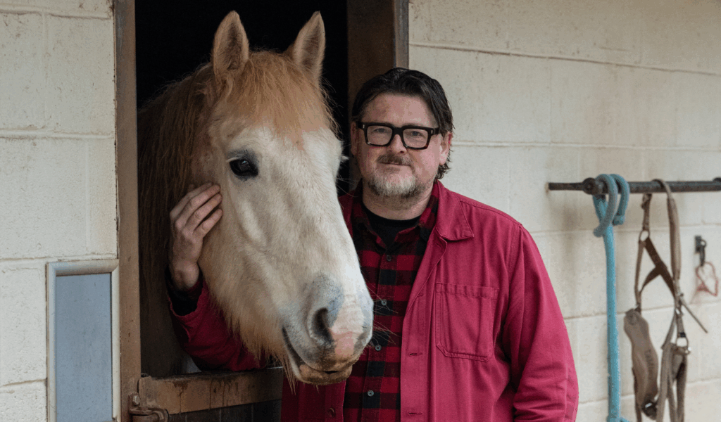 Rhodri Samuals, Chief Executive, wearing a red jacket and black checkered shirt, stands beside Vogue a grey horse in a stable doorway. The horse's head is leaning out of the stall, and the person has one hand resting gently on the horse's face. Behind them, the stable wall is visible with hanging tack and a blue rope.